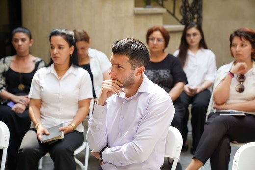 A man listens with his hand at his chin, thinking, while a woman beside him holds an open notebook, seated in a row of attentive colleagues at a bright outdoor courtyard session