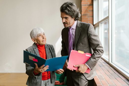 Two senior colleagues in business attire compare open folders of documents in a window-lit corridor