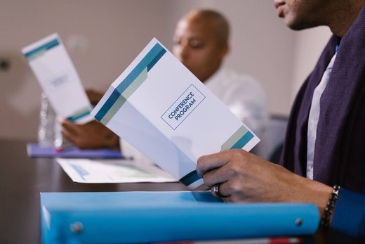 An attendee holds an open "Conference Program" folder while another delegate reads a program in the background at a shared table