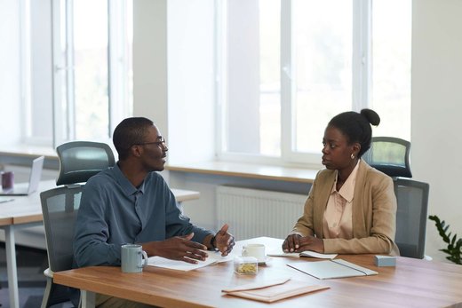 A man in a grey shirt gestures while talking with a woman in a tan blazer across a desk with open notebooks and coffee mugs, in a bright office with large windows