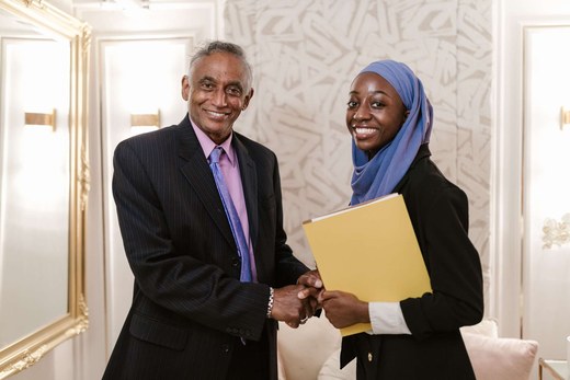 A handshake between two colleagues in business attire, a yellow folder held between them, bright indoor light