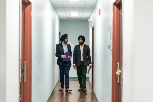 Two colleagues in suits and turbans walk together in conversation down an office corridor, each holding a folder