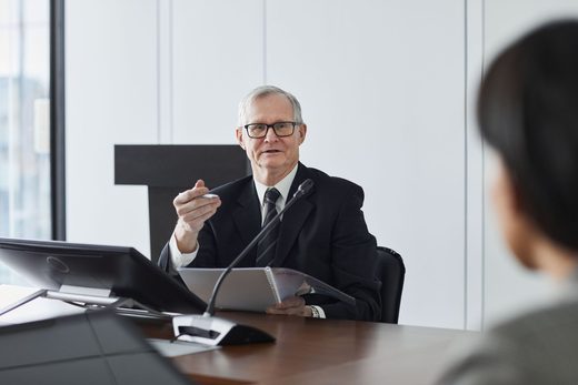 An older speaker in glasses gestures with a pen while speaking at a conference table, a monitor and spiral notebook in front of him, a colleague listening across the table