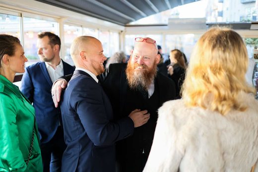 Two men embrace warmly in greeting amid a small group of colleagues, in a bright glass-walled reception space