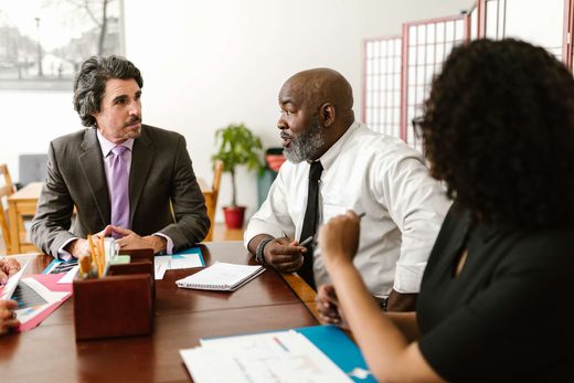 Three colleagues in business attire lean in mid-conversation at a table scattered with papers and folders