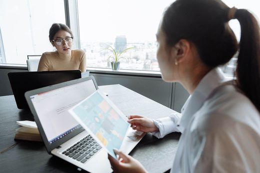 Two women sit across a table near a bright office window, one showing the other a printed colour infographic while open laptops sit on the table between them