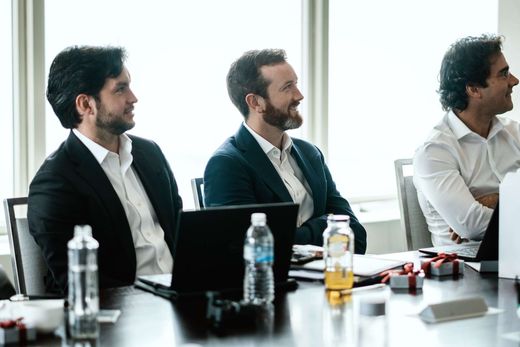 Three men in business attire sit at a table with laptops open and small ribbon-tied gift boxes, smiling as they listen to an unseen speaker in a bright, window-lit room
