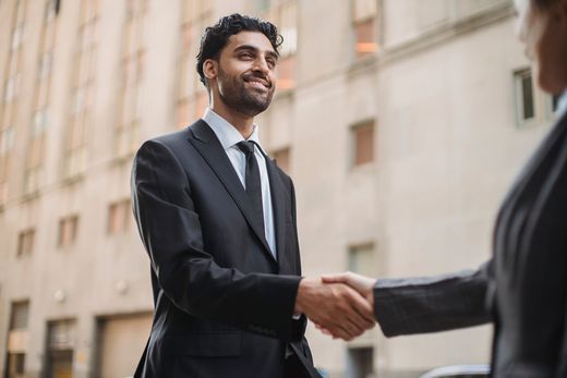 A man in a dark suit smiles while shaking hands with someone off-frame, a blurred city building facade behind him