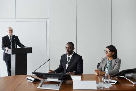 A seated panelist in a suit gestures while speaking at a conference table beside a colleague in a tweed blazer, with another presenter at a podium in the background