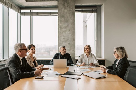 Five colleagues in business attire around a boardroom table with laptops and printed charts, city skyline visible through floor-to-ceiling windows