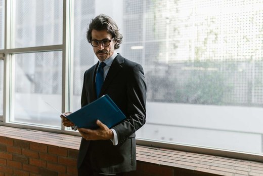 A man in a dark suit and glasses reads from a blue folder by a bright window, with city buildings visible outside