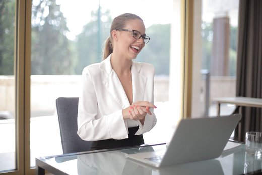 A woman in a white blazer and glasses laughs during a video call on a laptop at a glass desk in a bright, window-lined room