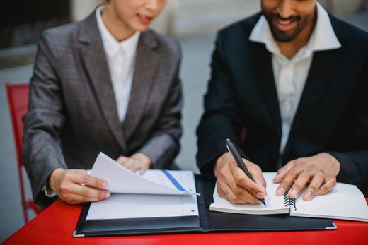 A man and a woman in business attire sit at an outdoor table, each writing in an open notebook or folder