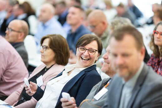 A badged conference delegate laughs mid-session, seated in a packed, brightly lit audience