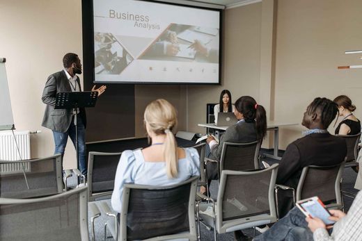 A presenter gestures while addressing a small seated group in a meeting room, a projection screen behind them