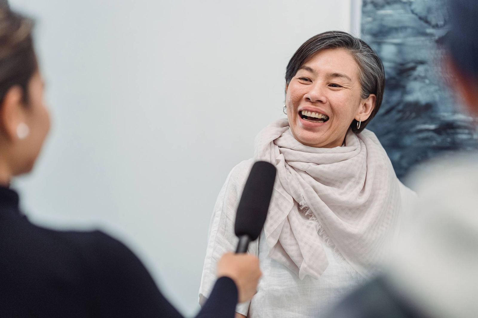 A woman in a cream scarf speaks animatedly while someone holds a handheld microphone toward her, against a bright white gallery wall