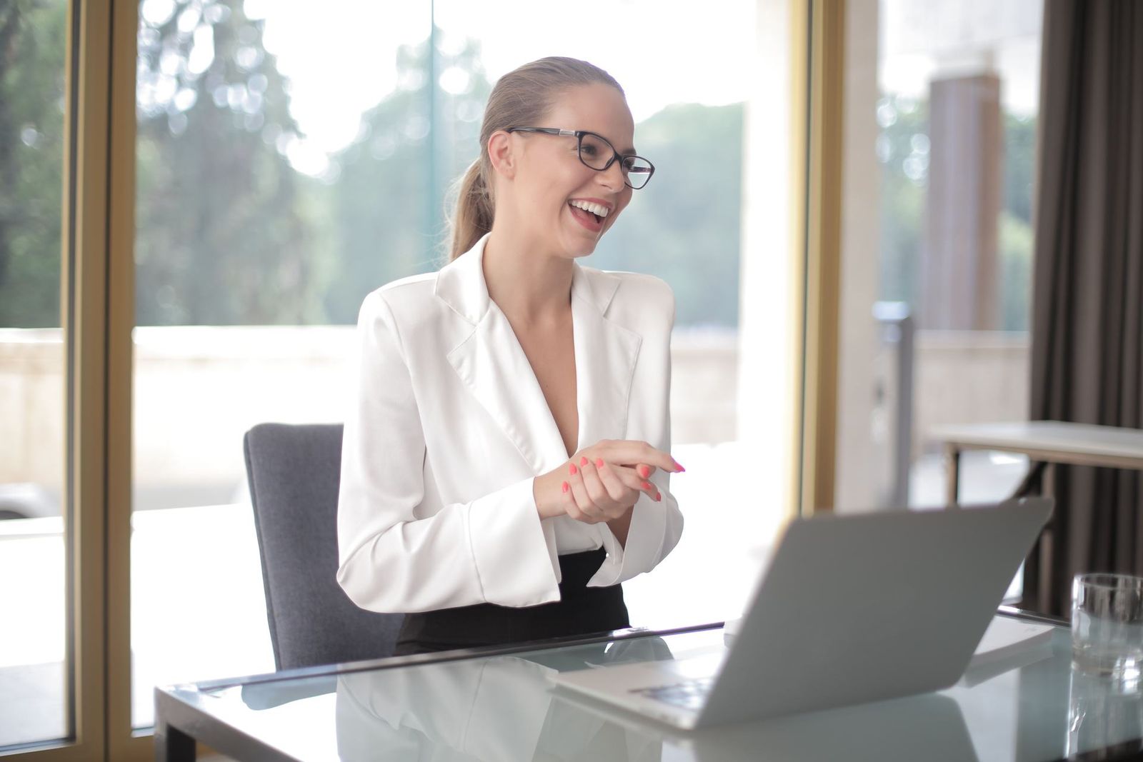 A woman in a white blazer and glasses laughs during a video call on a laptop at a glass desk in a bright, window-lined room