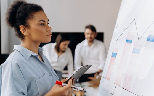A colleague presents a data chart on a whiteboard while two others review notes at a table behind her