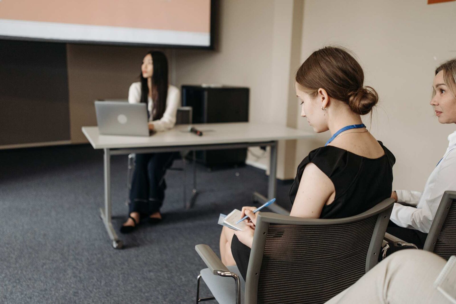 A delegate wearing a conference lanyard takes notes in the front row while a presenter speaks from a table