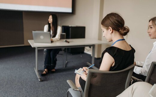 A delegate wearing a conference lanyard takes notes in the front row while a presenter speaks from a table