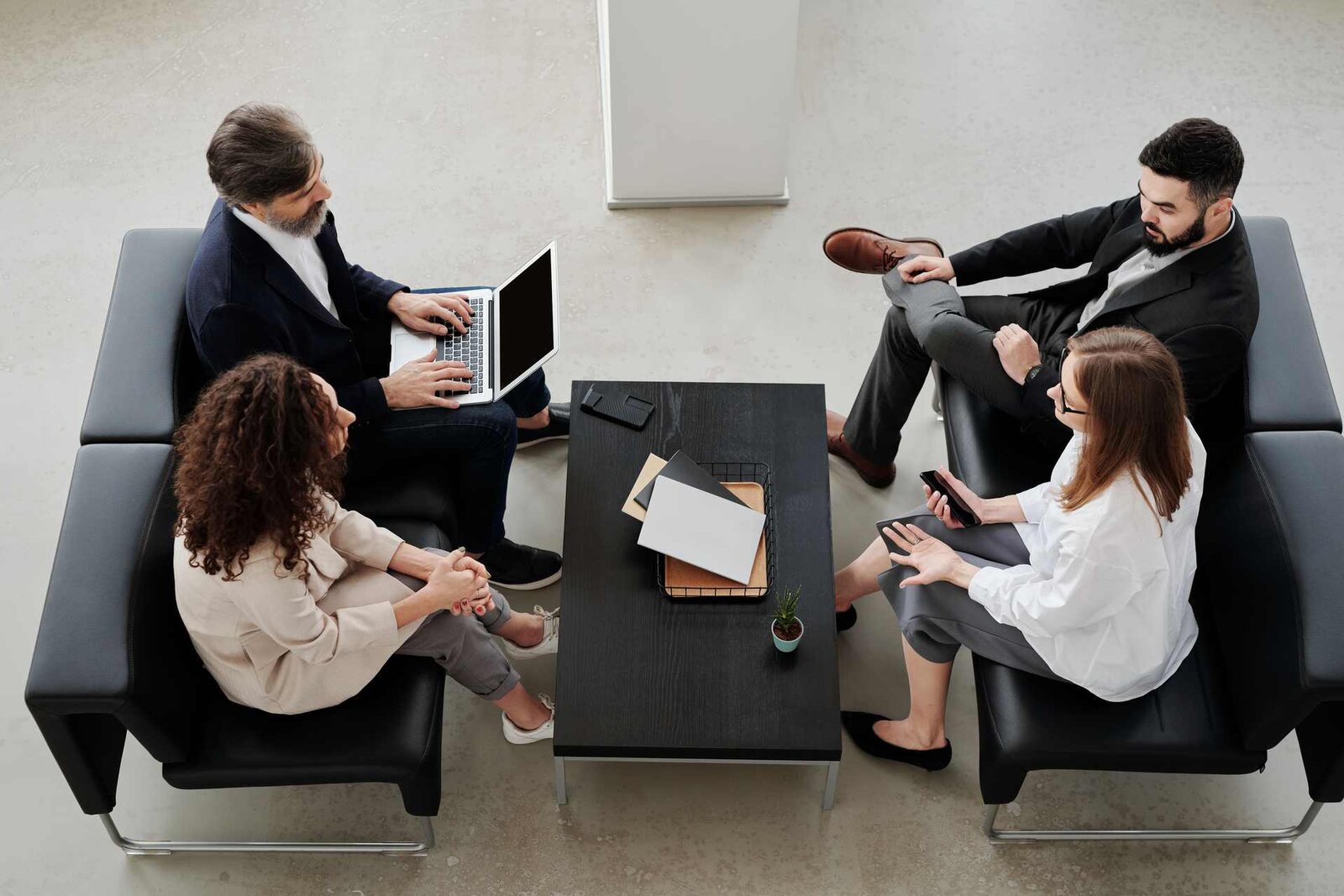 Four professionals in business attire sit around a coffee table in black lounge chairs, one working on a laptop, seen from overhead