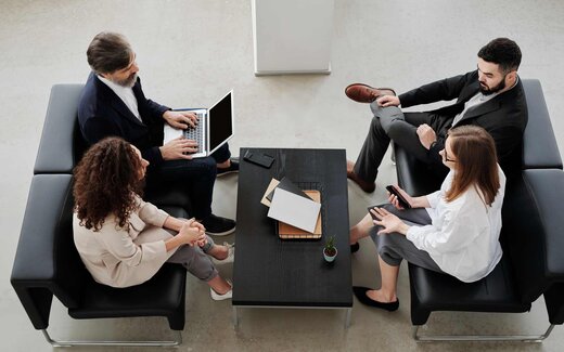 Four professionals in business attire sit around a coffee table in black lounge chairs, one working on a laptop, seen from overhead