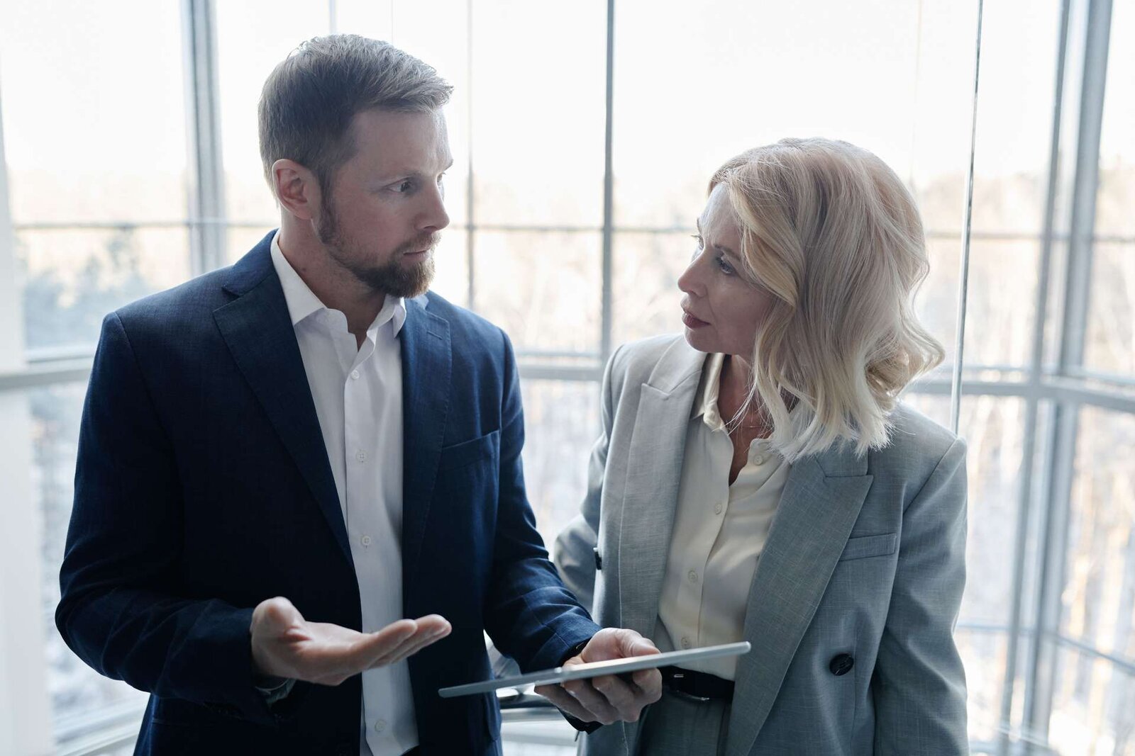 A man and a woman in business attire in serious discussion, one holding a tablet, in front of a bright curved glass wall