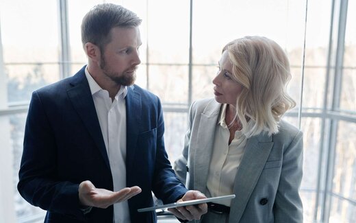 A man and a woman in business attire in serious discussion, one holding a tablet, in front of a bright curved glass wall