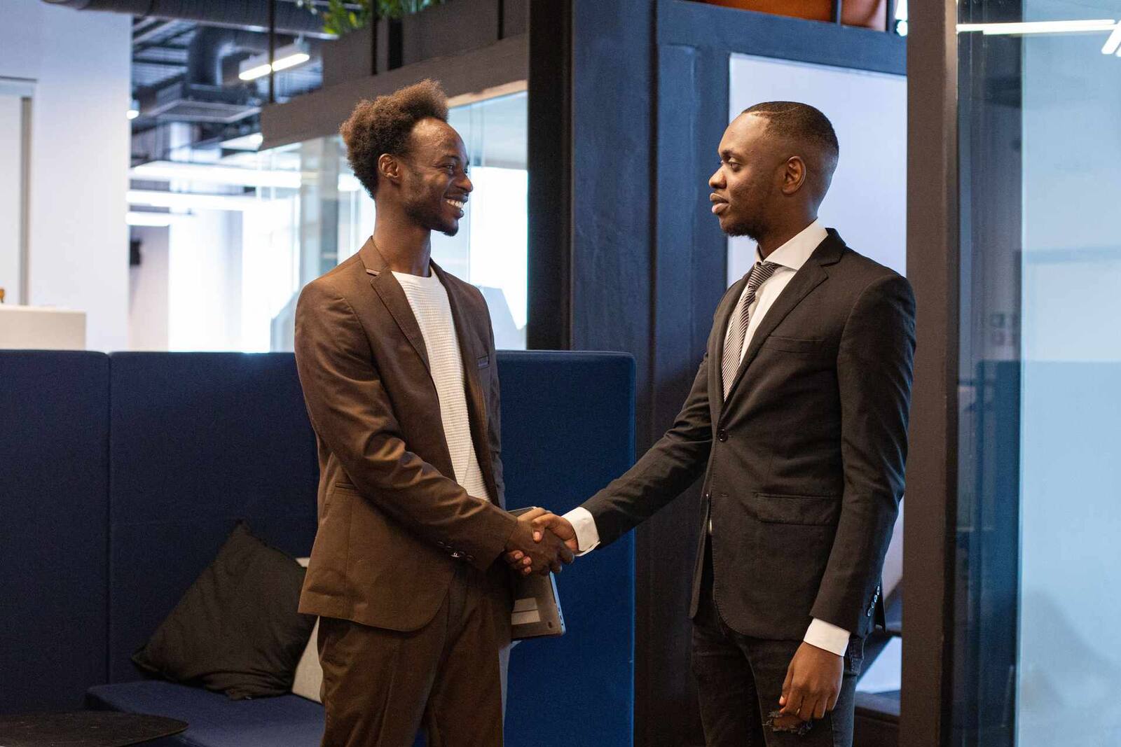 Two colleagues in business attire shake hands and smile in a bright glass-walled office lounge