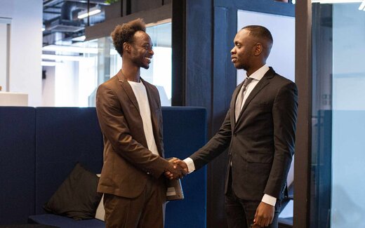 Two colleagues in business attire shake hands and smile in a bright glass-walled office lounge