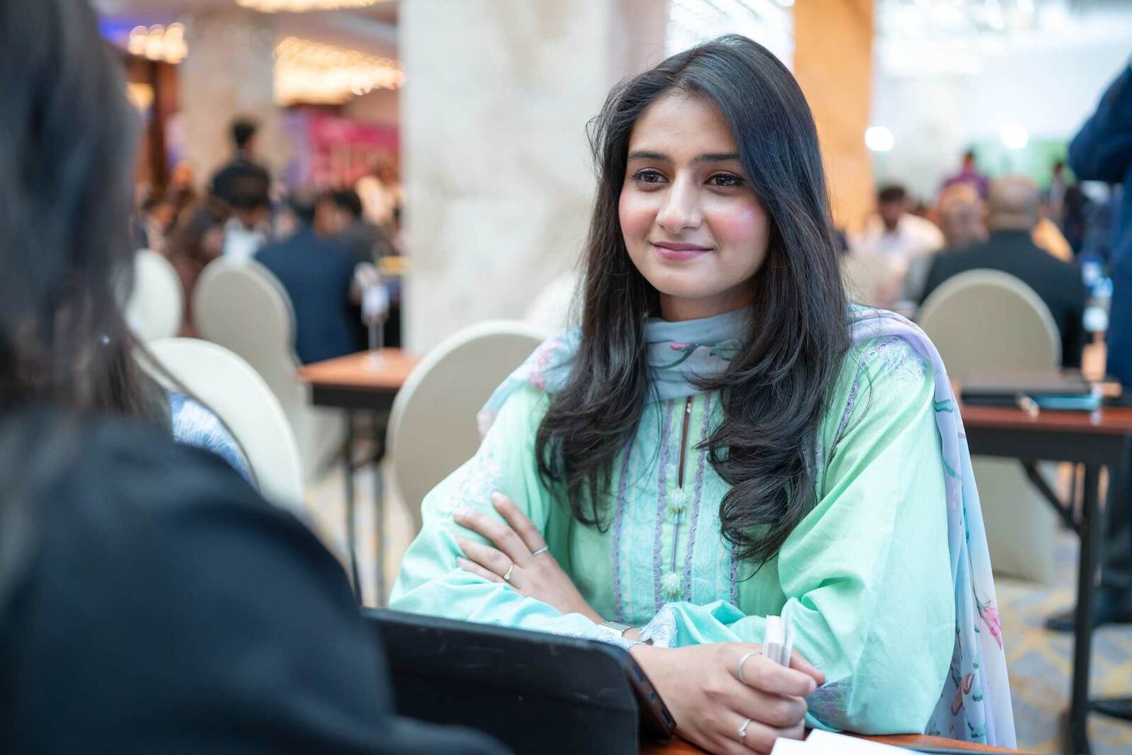 A delegate smiles mid-conversation at a partnering table during a busy conference ballroom concourse