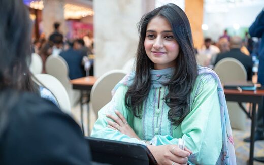 A delegate smiles mid-conversation at a partnering table during a busy conference ballroom concourse