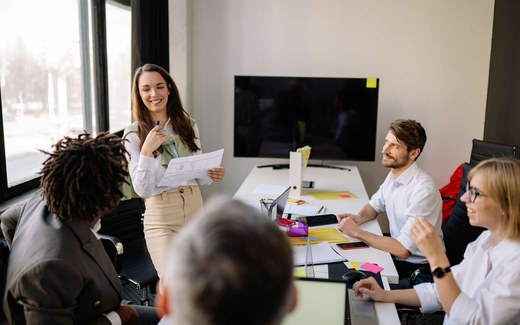 A presenter shares a printed chart with colleagues gathered around a bright meeting table