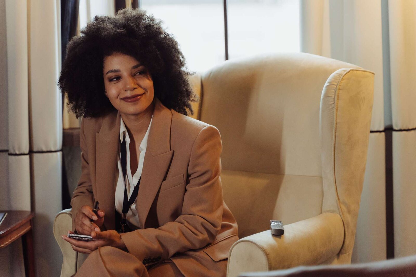 A woman in a tan suit with a lanyard and notebook, pen in hand, listening attentively in a hotel lounge chair