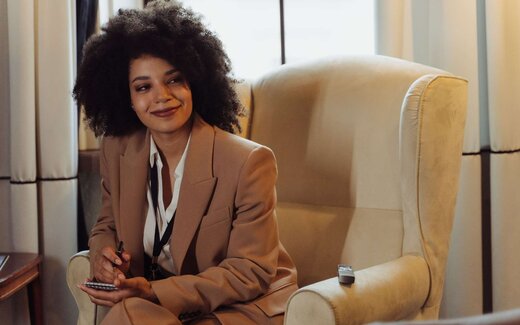 A woman in a tan suit with a lanyard and notebook, pen in hand, listening attentively in a hotel lounge chair