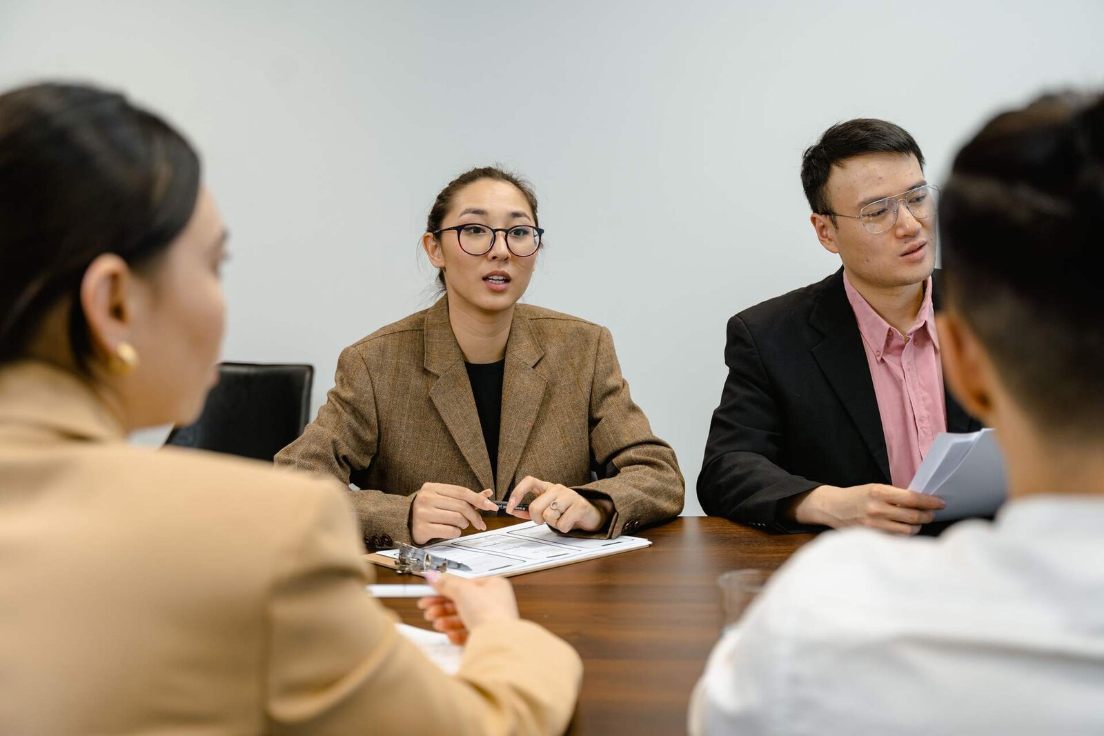 A woman in a brown blazer speaks across a meeting table to colleagues during a discussion