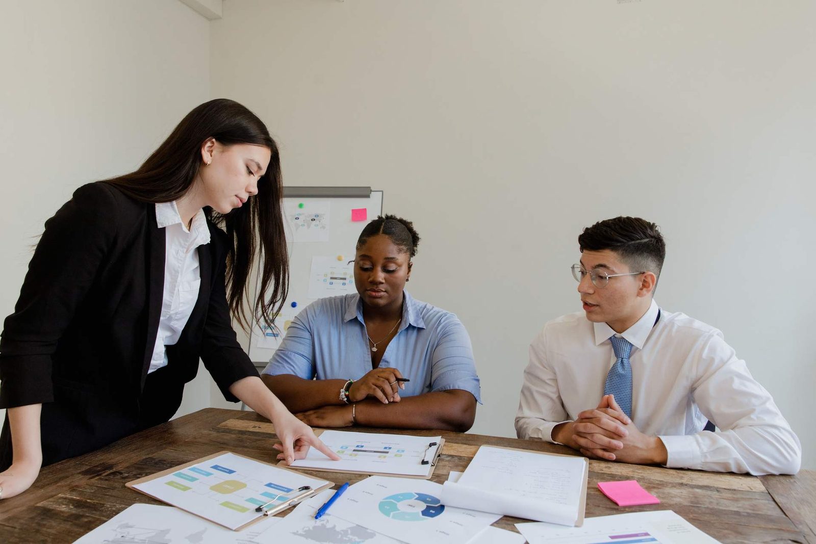 A presenter reviews printed process and data charts with two seated colleagues at a table