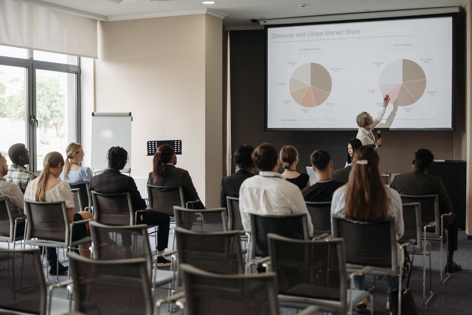 A presenter gestures toward a large projected slide while delegates look on from rows of seats in a bright session room