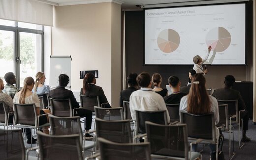 A presenter points at a projected slide before rows of seated delegates in a bright session room