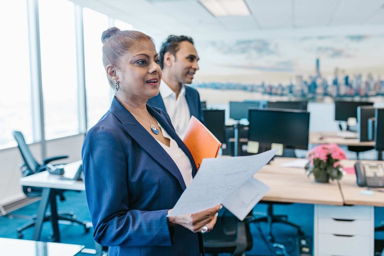 Two colleagues in navy blazers consult a printed document while standing in a bright, modern office