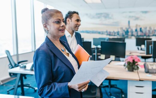 Two colleagues in navy blazers consult a printed document while standing in a bright, modern office