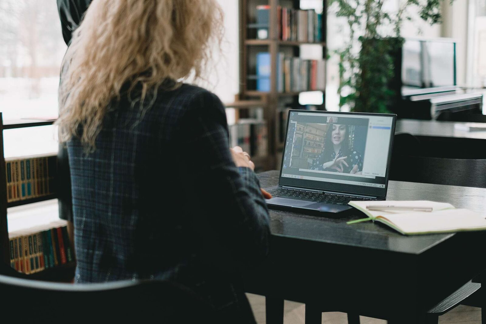 A woman seen from behind watches a recorded presenter on a laptop at a desk in a bright library
