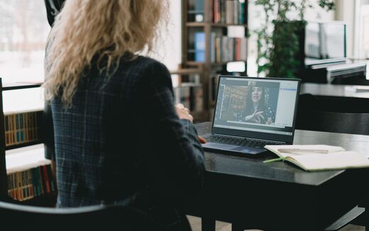 A woman seen from behind watches a recorded presenter on a laptop at a desk in a bright library