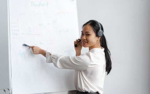 A smiling presenter wearing a headset microphone points at a hand-drawn product roadmap on a flipchart