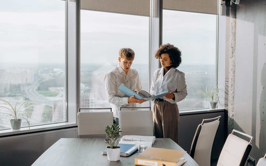 Two colleagues stand at an office window reviewing a document together, one holding a pen