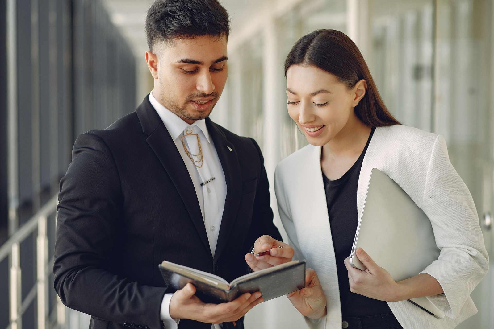 Two colleagues stand in a bright corridor reviewing a notebook together, one holding a laptop