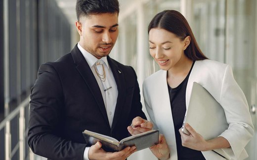Two colleagues stand in a bright corridor reviewing a notebook together, one holding a laptop