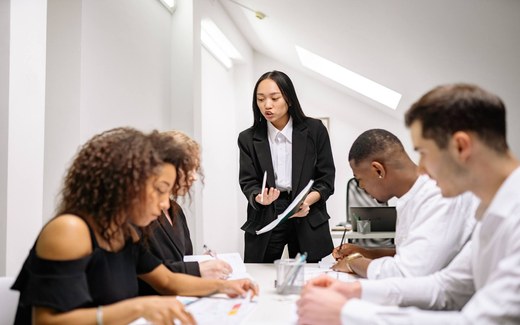 A presenter briefs colleagues seated around a table, taking notes, in a bright office