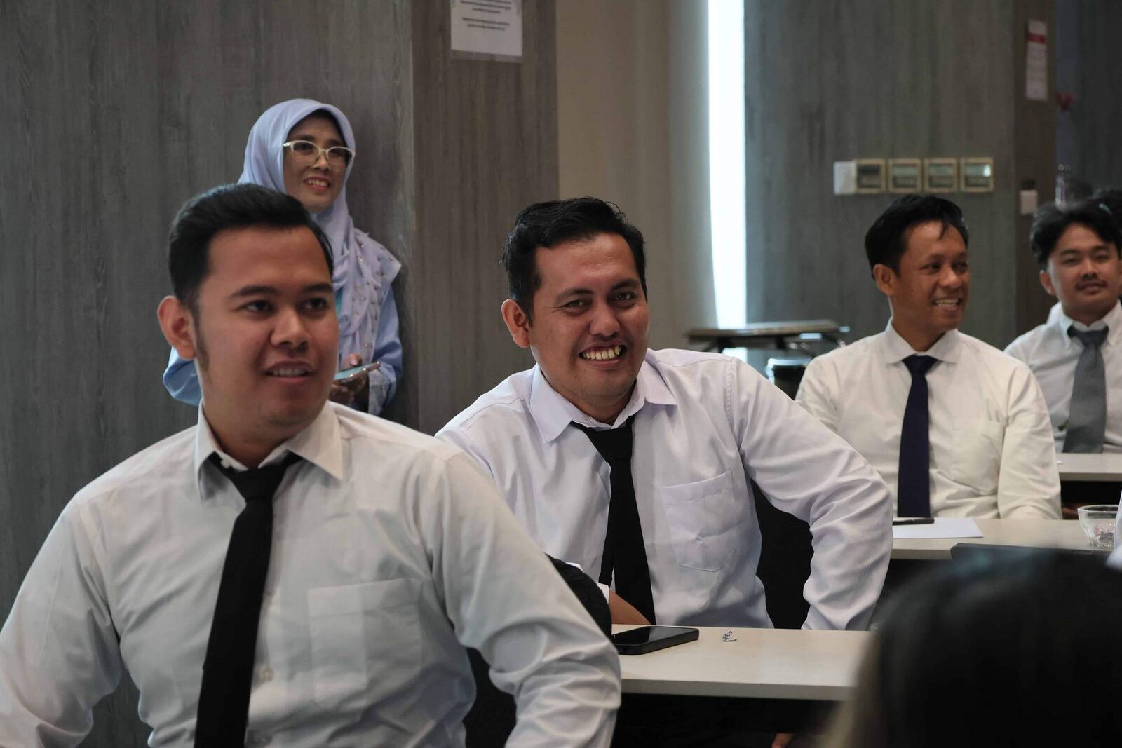 Men in white shirts and ties seated at training tables, smiling toward an unseen speaker, with a woman in a light-blue hijab standing and smiling at the back of the room
