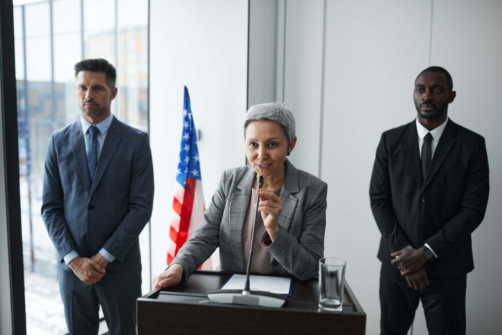 A speaker addresses the room from a podium beside a US flag, flanked by two colleagues